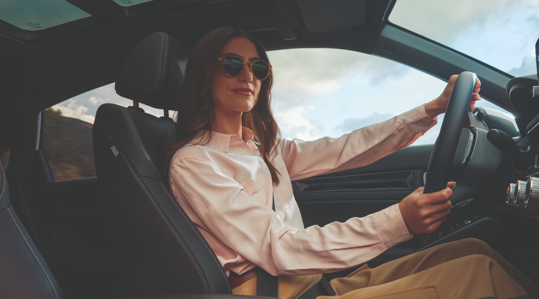 A person wearing sunglasses and a light pink shirt drives a car with a modern interior. The driver's seat has black leather upholstery, and the steering wheel is visible. Through the windshield, a cloudy sky can be seen.